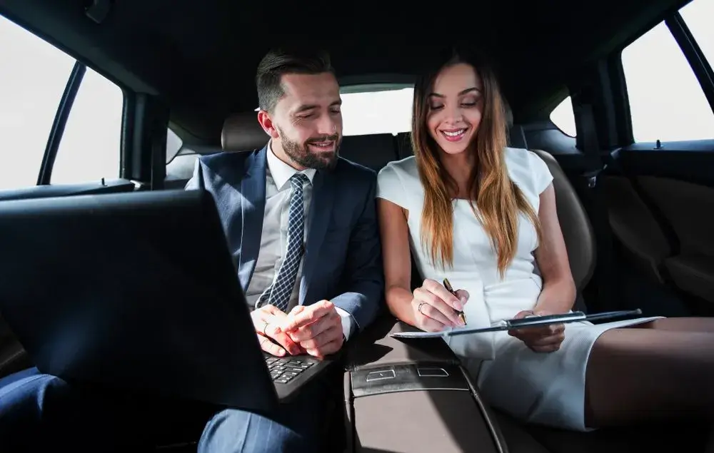 City-To-City Transfers A man and a woman dressed in business attire sit in the back seat of a limousine, smiling while reviewing documents together on a clipboard and laptop