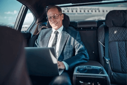 A professional businessman in a suit and glasses smiling while working on a laptop in the backseat of a luxury black car, representing Gateway Limousine's executive transportation and St. Louis airport car services.