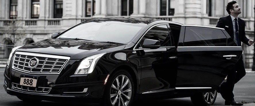 A professional chauffeur in a black suit standing by the open rear door of a black Cadillac luxury sedan in downtown St. Louis, demonstrating the premium service provided by Gateway Limousine.