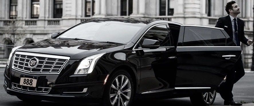 Our Blogs 8 A professional chauffeur in a black suit standing by the open rear door of a black Cadillac luxury sedan in downtown St. Louis, demonstrating the premium service provided by Gateway Limousine.